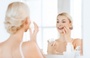 beauty, skin care and people concept - smiling young woman applying cream to face and looking to mirror at home bathroom