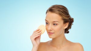 beauty, people and skincare concept - young woman cleaning face with exfoliating sponge over blue background